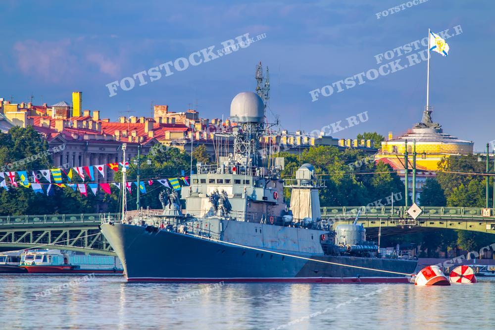 На фотографии изображен военный корабль, стоящий на якоре на фоне исторической застройки Санкт-Петербурга. Видны здания набережной, украшенные флагами, и Андреевский флаг на корабле. Фото может использоваться для туристических буклетов, открыток и рекламных материалов.